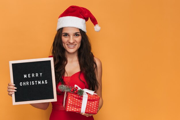 Une femme caucasienne souriante dans un bonnet de Noel rouge tenant une boîte-cadeau et un tableau à lettres avec l'inscription Joyeux Noël dessus. Sur un fond isolé jaune. Une place pour votre texte.