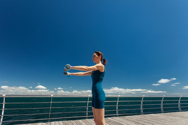 Une femme caucasienne sportive faisant de l'exercice avec des haltères sur le remblai par une belle journée ensoleillée avec un ciel bleu et des nuages Concept de style de vie sain Grand angle