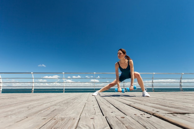 Une femme caucasienne sportive faisant de l'exercice avec des haltères sur le remblai par une belle journée ensoleillée avec un ciel bleu et des nuages Concept de style de vie sain Grand angle