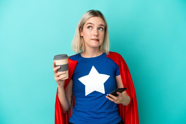 Femme caucasienne de super héros isolée sur fond bleu tenant du café à emporter et un mobile tout en pensant à quelque chose