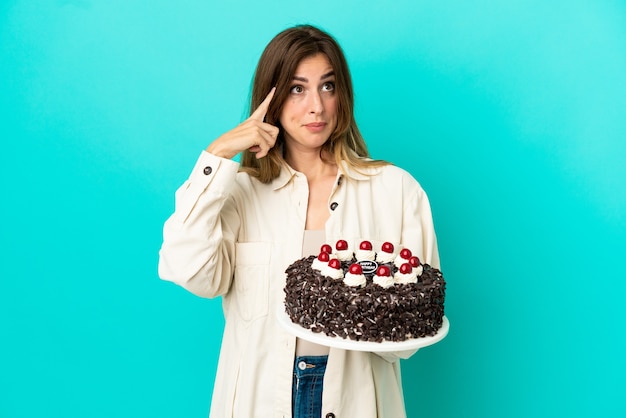 Femme caucasienne tenant un gâteau d'anniversaire isolé sur fond bleu ayant des doutes et pensant