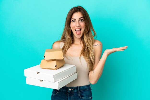 Femme caucasienne tenant des pizzas et un hamburger isolé sur fond bleu avec une expression faciale choquée