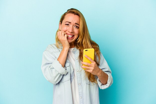 Femme caucasienne tenant un téléphone portable isolé sur fond bleu se ronger les ongles, nerveuse et très anxieuse.