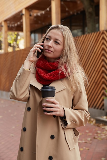 Femme caucasienne en trench-coat beige appelant par smartphone avec une tasse de café