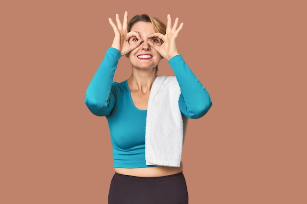 Femme caucasienne en vêtements de sport avec une serviette montrant le signe OK sur les yeux