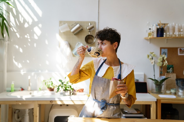 Une femme céramiste boit du café et mange une collation dans un studio d'art à la pause de la classe de maître produit de la poterie