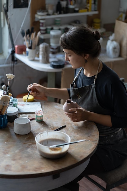 Une femme céramiste concentrée travaillant dans un atelier est assise à table et décore la poterie avec des peintures