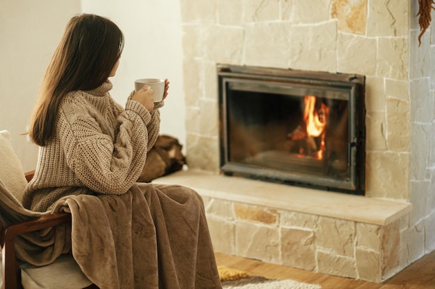 Femme en chandail confortable tenant une tasse de thé chaud à la cheminée automne hygge Chauffage maison avec poêle