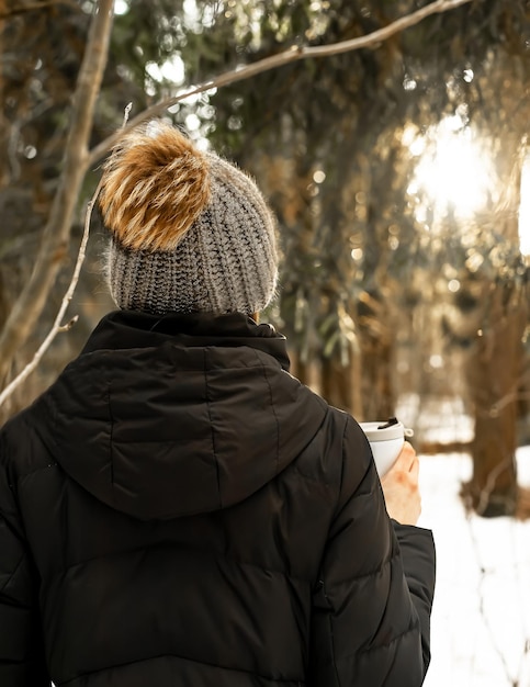 Femme en chapeau de laine tricoté avec pompom tenant une tasse thermo dans la nature hivernale