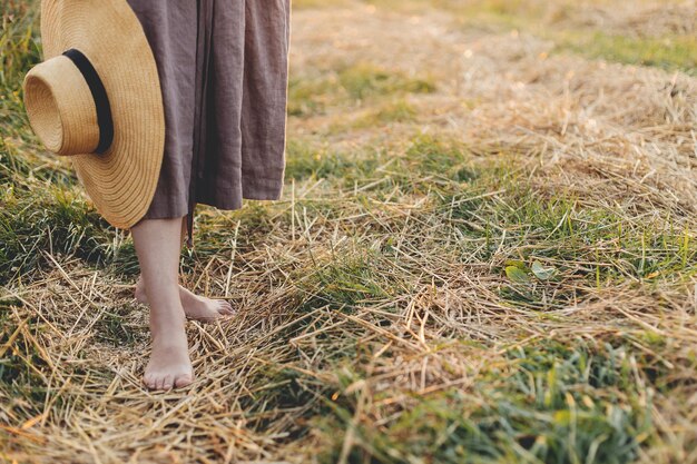 Femme avec chapeau à la main marchant pieds nus sur un champ de paille dans la lumière du coucher du soleil gros plan Vie lente rurale Moment tranquille atmosphérique Jeune femme en robe de lin rustique relaxante dans la campagne estivale