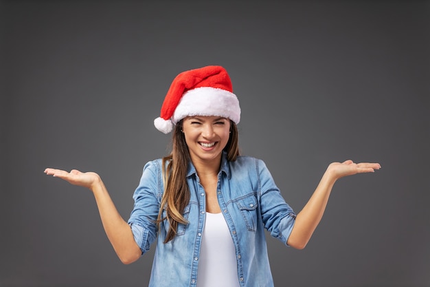 Femme avec chapeau de père Noël habillé décontracté tenant les mains en l'air comme si elle tenait quelque chose.