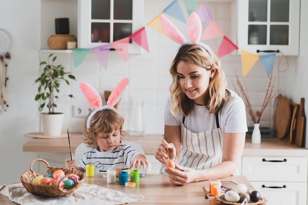 Une femme charmante et son fils de deux ans dans des oreilles de lapin peignent des oeufs de Pâques avec des peintures assis à la table de la cuisine