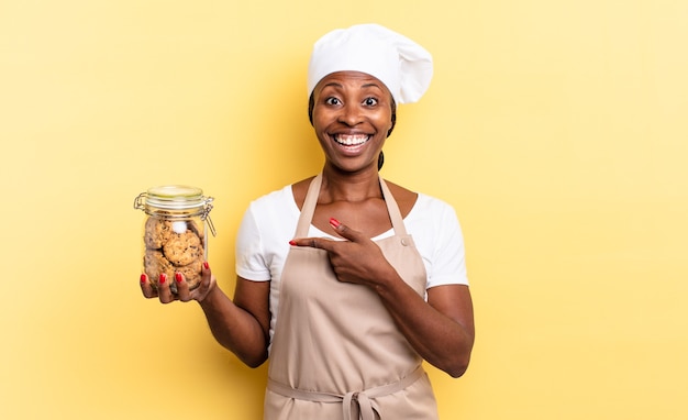 Femme Chef Afro Noire à L'air Excitée Et Surprise Pointant Sur Le Côté Et Vers Le Haut Pour Copier L'espace. Notion De Cookies