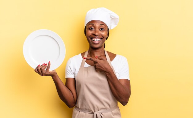 Femme chef afro noire à l'air excitée et surprise pointant vers le côté et vers le haut pour copier l'espace. concept d'assiette vide