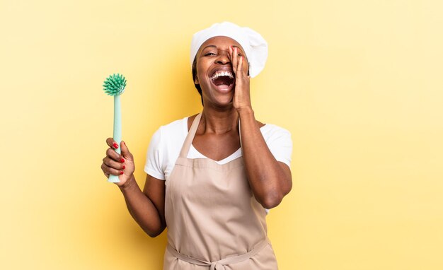 Femme chef afro noire se sentant heureuse, excitée et positive, donnant un grand cri avec les mains à côté de la bouche, appelant. concept de nettoyage de la vaisselle