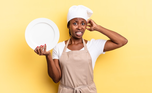 Femme chef afro noire se sentant stressée, inquiète, anxieuse ou effrayée, les mains sur la tête, paniquée par erreur. concept d'assiette vide