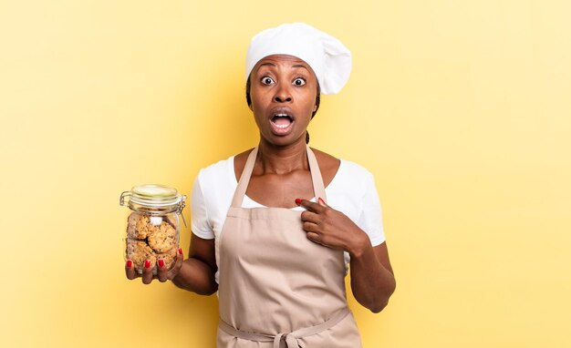 Femme chef afro noire semblant choquée et surprise avec la bouche grande ouverte, pointant vers soi. notion de cookies