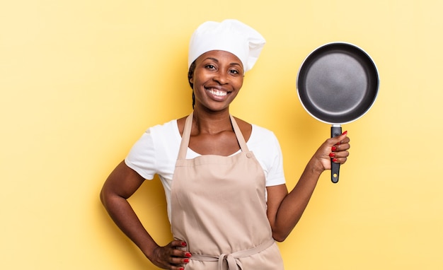 Femme chef afro noire souriant joyeusement avec une main sur la hanche et une attitude confiante, positive, fière et amicale