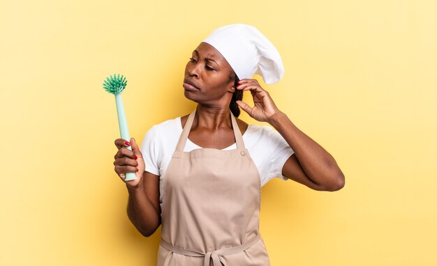 Femme chef afro noire souriant joyeusement et rêvant ou doutant, regardant sur le côté. concept de nettoyage de la vaisselle