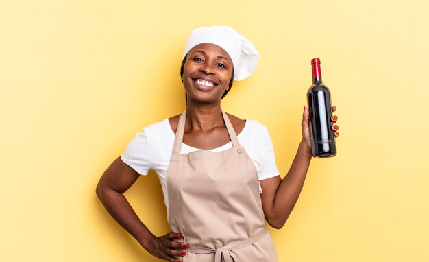 Femme chef afro noire souriante joyeusement avec une main sur la hanche et une attitude confiante, positive, fière et amicale. concept de bouteille de vin