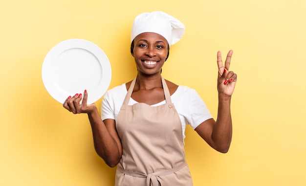 Femme chef afro noire souriante et semblant amicale, montrant le numéro deux ou la seconde avec la main en avant, comptant à rebours. concept d'assiette vide