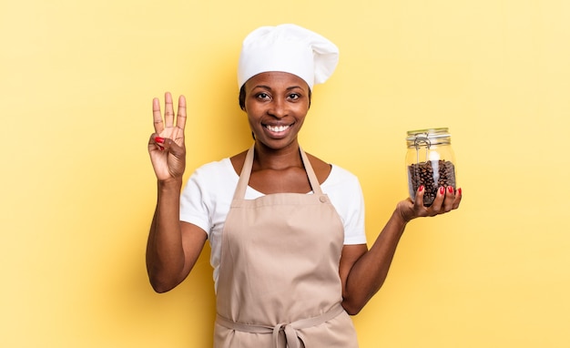 Femme chef afro noire souriante et semblant amicale, montrant le numéro trois ou troisième avec la main en avant, compte à rebours. concept de grains de café