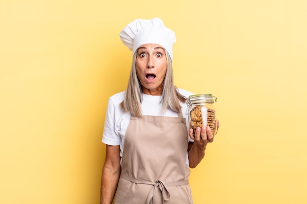 Femme chef d'âge moyen ayant l'air très choquée ou surprise, regardant la bouche ouverte en disant wow avec des cookies