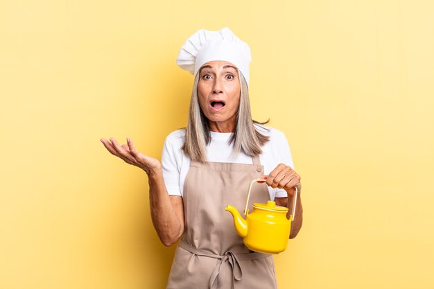 Femme chef d'âge moyen bouche bée et étonnée, choquée et étonnée d'une surprise incroyable et tenant une théière