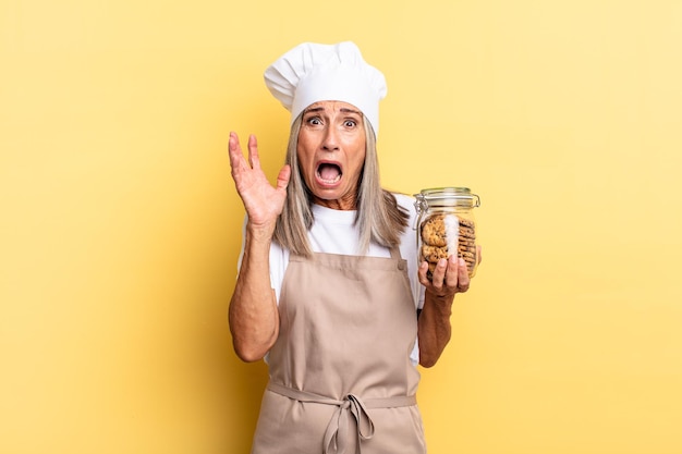 Femme chef d'âge moyen criant avec les mains en l'air, se sentant furieuse, frustrée, stressée et contrariée par les cookies