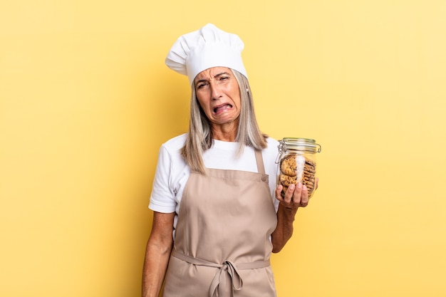 Femme chef d'âge moyen se sentant perplexe et confuse, avec une expression stupide et abasourdie en regardant quelque chose d'inattendu avec des cookies