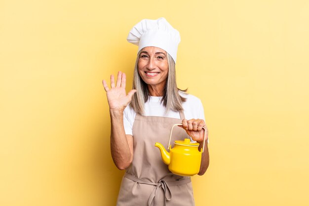 Femme chef d'âge moyen souriant joyeusement et joyeusement, agitant la main, vous accueillant et vous saluant, ou disant au revoir et tenant une théière