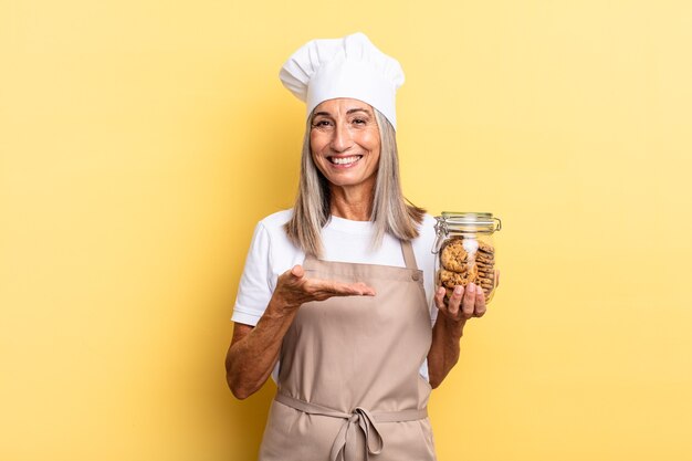 Femme chef d'âge moyen souriant joyeusement, se sentant heureuse et montrant un concept dans l'espace de copie avec la paume de la main avec des cookies