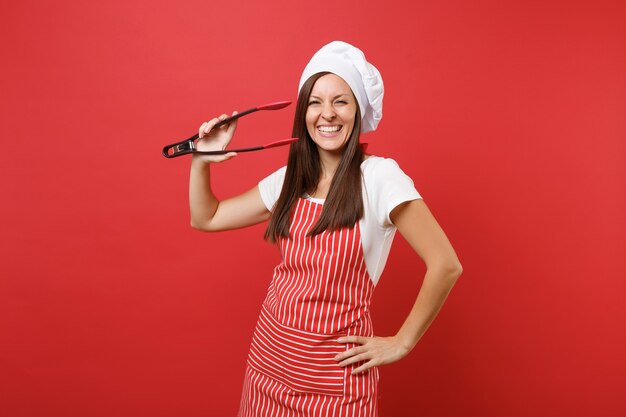 Femme chef cuisinier ou boulanger en tablier rayé, t-shirt blanc toque chapeau de chef isolé sur fond de mur rouge. Une femme tient une salade de cuisine servant une paire de pinces en plastique pour griller. Maquette du concept d'espace de copie.