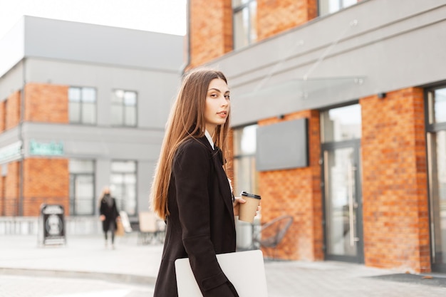 Femme chef d'entreprise professionnelle avec des vêtements élégants noirs avec ordinateur portable et tasse de café se promène dans la ville