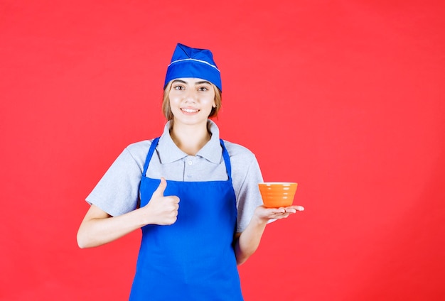 Femme chef en tablier bleu tenant une tasse de nouilles et montrant le pouce vers le haut