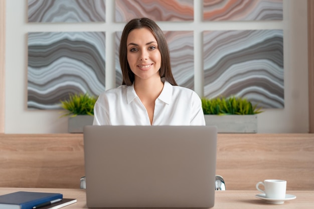 Femme en chemise blanche beau sourire assis à table avec ordinateur portable à l'intérieur du bureau en bois