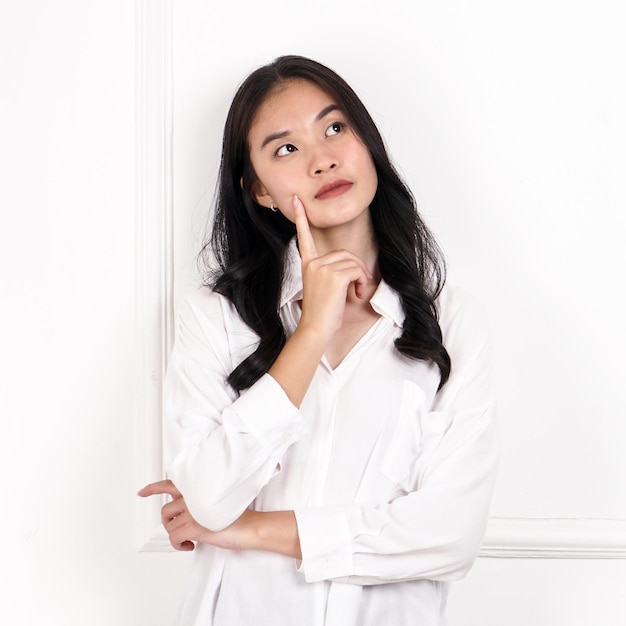 Une femme en chemise blanche est debout contre un mur blanc et a sa main sur son menton.