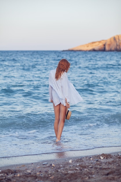 Une femme en chemise blanche se tient dans l'eau à la plage et regarde la mer.