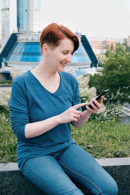 Femme en chemise bleue assise dans un quartier d'affaires et regarde l'écran d'un smartphone