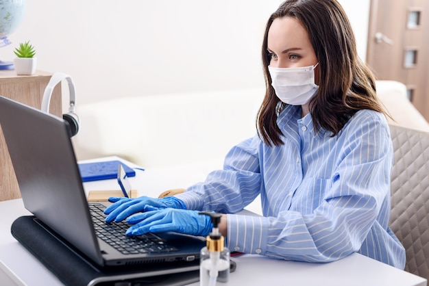 Femme En Chemise Bleue Avec Masque Médical Sur Le Visage Et Des Gants Médicaux Jetables Se Trouve à L'ordinateur Portable, Travail à Distance à La Maison Sous Le Concept De Quarantaine