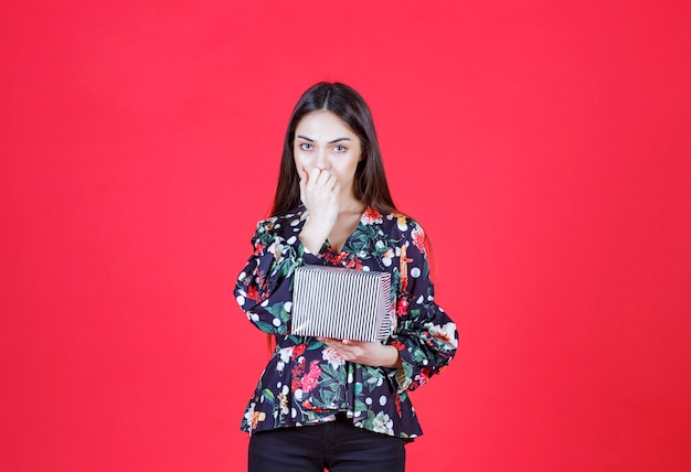 femme en chemise florale tenant une boîte-cadeau en argent et a l'air réfléchie.