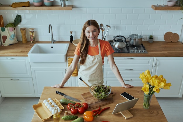 une femme en chemise orange se tient devant un comptoir de cuisine avec des légumes et des fruits