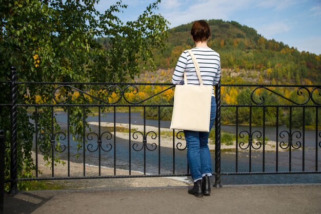 Femme en chemise rayée à manches longues portant un sac à provisions réutilisable vide près de la maquette de la balustrade du pont