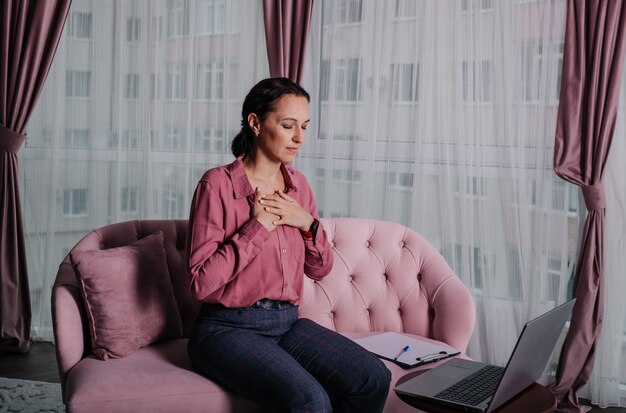 Une femme en chemise rose est assise sur le canapé et effectue une consultation à distance à l'aide d'un ordinateur portable