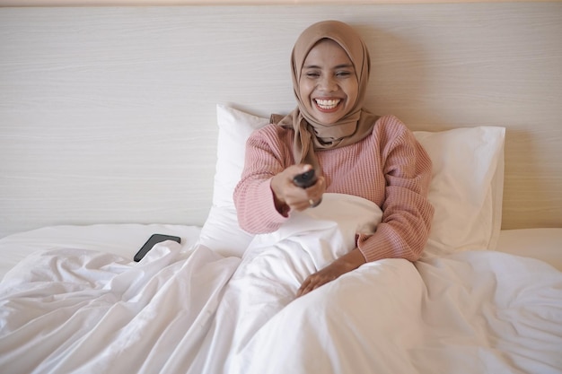 Une femme en chemise rose est assise sur un lit et regarde la télévision