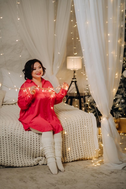 Une femme en chemise rouge dans une chambre décorée pour Noël est assise sur un lit Ambiance Nouvel An Accueil