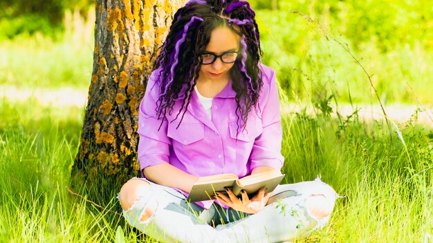 Une femme en chemise violette et lunettes lit une littérature sous un arbre Une fille se repose avec un livre dans la forêt Une femme avec un livre est assise sur l'herbe par une claire journée d'été