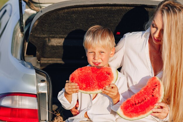 Femme cheveux assez blonds avec petit fils blond au coucher du soleil se détendre derrière la voiture et manger la pastèque. été, voyages, nature et grand air à la campagne.