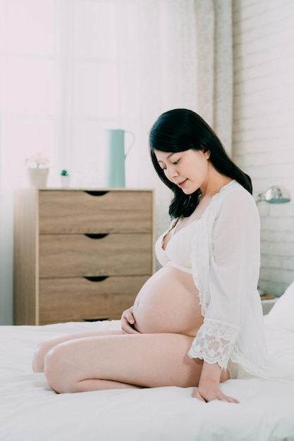 femme chinoise asiatique de mode sexy regarde le ventre de femme enceinte nu assis dans un lit de confort blanc. jeune maman de maternité se détendant dans la chambre tandis qu'une main tenant le ventre nu attention à prendre soin du bébé à naître