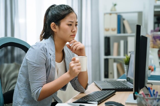 Femme chinoise asiatique pensive en smart casual avec ordinateur de bureau regardant l'écran tenant une tasse de café au bureau à domicile. jeune employée réfléchie assise au pc remue-méninges pendant boire du thé.